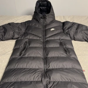 Svart lång pufferjacka från Nike Storlek S - Säljer en svart pufferjacka från Nike med huva och två praktiska fickor med dragkedja. Jackan har en lång passform och är perfekt för kalla dagar. Ikonisk Nike-logga på bröstet och snygga breda paneler ger en modern look.