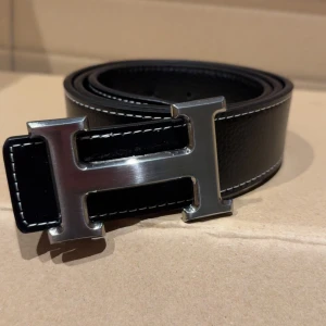 Svart skärp med H-spänne - Snyggt svart bälte med ett ikoniskt H-format metallspänne i silver med vit kontrastsöm längs kanterna, vilket ger en stilren och lyxig känsla. Perfekt accessoar för att lyfta din outfit