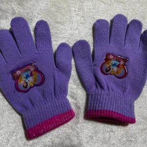 Lila stickade prinsesshandskar - Säljer ett par lila stickade handskar med rosa muddar och motiv av Disneyprinsessor på ovansidan. Perfekt för dig som gillar färgglada accessoarer och vill ha något extra gulligt till vintern.
