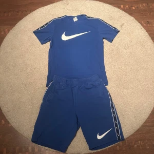 Blå Nike shorts med vita detaljer - Säljer ett par blå Nike t-shirt och shorts med vit logga framtill och vita detaljer längs sidorna. Shortsens midja är elastisk och de har fickor på båda sidor. Perfekta för träning eller chill dagar. Materialet känns mjukt och sportigt.