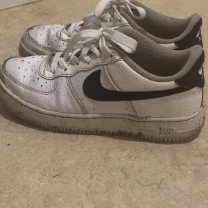 Nike Air Force 1 vita sneakers - Säljer ett par klassiska Nike Air Force 1 sneakers i vitt skinn med svart swoosh på sidan. Skorna har rund tå, snörning och platt sula. Perfekt för dig som gillar streetstyle och vill ha en ikonisk sneaker i garderoben. Tvättas inann dem skickas 