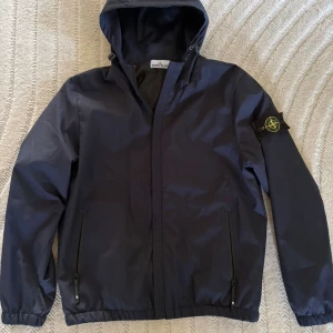 Mörkblå vindjacka från Stone Island - Snygg mörkblå vindjacka från Stone Island med huva och klassisk logotyp på vänster ärm. Jackan har dragkedja framtill, två fickor med dragkedja och elastiska muddar vid ärmslut och nederkant. Perfekt för dig som gillar streetwear och stilren design.