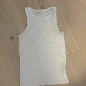 Vit ribbad linnetopp från H&M - En klassisk vit ribbad linnetopp från H&M. Toppen är ärmlös med rund halsringning och har en enkel, stilren design som passar till allt. Perfekt att matcha med jeans eller shorts för en avslappnad look.