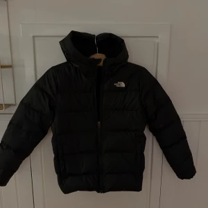Svart pufferjacka från The North Face - Svart pufferjacka från The North Face med fluffigt teddyfoder i huvan. Jackan har en hög krage, dragkedja framtill och är designad för att hålla dig varm under kalla dagar. Perfekt för dig som gillar stilren och funktionell ytterkläder. I storlek L men junior storlek så lite mindre. Köpt för cirka 2500kr