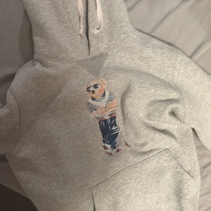 Grå hoodie Polo Ralph Lauren M - Säljer en grå hoodie från Polo Ralph Lauren i storlek M. Tröjan har en ikonisk björn med solglasögon tryckt på bröstet, stor huva med snörning och en klassisk känguruficka framtill. Perfekt för dig som gillar streetstyle och vill ha något bekvämt och snyggt.