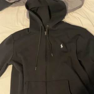 Svart zip från Polo Ralph Lauren - Svart hoodie från Polo Ralph Lauren med klassisk vit broderad logga på bröstet. Tröjan har dragkedja framtill, snörning i huvan och känguruficka. Tillverkad i mjuk bomullsmix, perfekt för chill dagar eller när du vill ha en clean streetstyle.