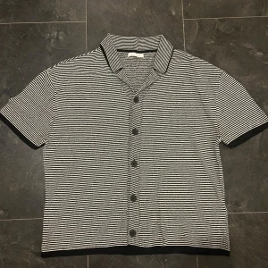 Randig svartvit kortärmad skjorta - Snygg kortärmad skjorta med svartvita tunna ränder och klassisk krage. Skjortan har svarta knappar framtill och kontrastkant vid ärmslut och nederkant. Materialet känns mjukt och har en stickad look, perfekt för dig som gillar retrovibbar och stilrena detaljer.