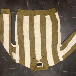 Randig stickad tröja från Pull&Bear - Snygg stickad tröja från Pull&Bear med breda olivgröna och beige ränder. Tröjan har en klassisk rund halsringning och långa ärmar. Perfekt för dig som gillar retrovibbar och vill ha en mysig, unik look. Ribbstickade muddar vid hals, ärmslut och nederkant.