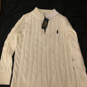 Vit kabelstickad tröja Polo Ralph Lauren - Kabelstickad vit tröja från Polo Ralph Lauren i storlek S. Tröjan har ribbad krage, muddar och nederkant, samt en half zip-dragkedja vid halsen. Klassisk broderad logga på bröstet och mjukt material som känns lyxigt. Perfekt för dig som gillar stilrena och tidlösa plagg.