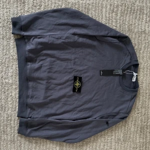 Grå sweatshirt från Stone Island - Säljer en stilren grå sweatshirt från Stone Island med klassisk logotyp på ärmen. Tröjan har rund halsringning och långa ärmar, perfekt för en avslappnad look. Tillverkad i mjuk bomull för skön komfort.