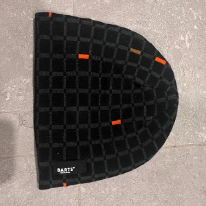 Svart rutmönstrad mössa från Barts - Svart mössa från Barts med rutmönster i mörkgrått och inslag av orange och bruna detaljer. Snygg och stilren design som sticker ut med sina färgklickar. Mjuk insida i fleece för extra komfort och värme. Perfekt för kalla dagar.