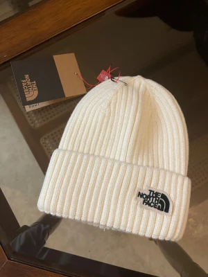 Vit ribbad mössa från The North Face - Snygg vit mössa från The North Face med ribbad design och uppvikt kant. Tillverkad i 100% bomull, perfekt för att hålla dig varm och stilren under kyliga dagar. Logotypen är broderad i svart på framsidan för en clean look.