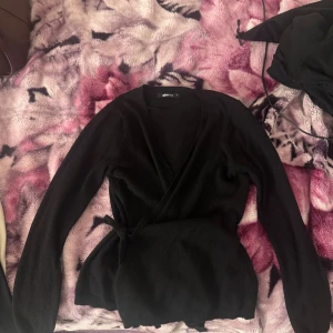 Svart omlottkofta från Gina Tricot - Svart omlottkofta från Gina Tricot med långa ärmar och knyt i midjan. Mjuk stickad känsla som sitter snyggt och ger en clean look. Perfekt för dig som gillar stilrena plagg med lite extra detalj. 