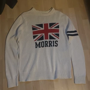 Vit stickad tröja Morris med flagga - Snygg vit stickad tröja från Morris i storlek M. Stor Union Jack-flagga och Morris-tryck på bröstet, samt två mörkblå ränder på ena ärmen. Rund hals och ribbade muddar vid ärmslut och nederkant. Perfekt för dig som gillar klassisk stil med en twist.