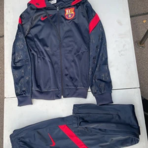 Barcelona Nike träningsset marinblå - Barcelona träningsset från Nike i marinblått med röda detaljer. Jackan har huva, dragkedja och klubbmärke på bröstet samt mönstrade ärmar med klubbens logga. Byxorna har elastisk midja och röd Nike-logga. Perfekt för dig som älskar fotboll och vill ha en snygg look.