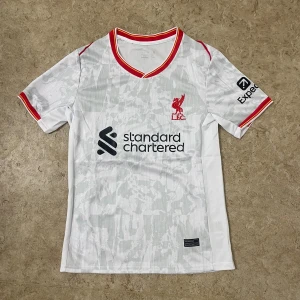 Liverpool vit fotbollströja Nike - Säljer en officiell Liverpool fotbollströja från Nike i vit med ljusgrått mönster. Tröjan har korta ärmar och röd-gula detaljer vid krage och ärmslut. Materialet är lätt och andas, perfekt för match eller träning. Tryck med Standard Chartered och Liverpool FC-logga på bröstet.