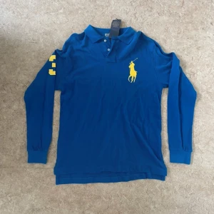 Blå långärmad pikétröja Polo Ralph Lauren - Blå långärmad pikétröja från Polo Ralph Lauren med stor gul logga på bröstet och gula siffror på ena ärmen. Klassisk krage med två knappar och ribbade muddar. Tröjan är i bomull och har en snygg custom fit-passform.