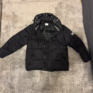 Burberry jacka storlek S - Helt ny, Svart burberry pufferjacka med huva och rutigt foder. Jackan har dragkedja och tryckknappar framtill, två sidofickor och en patch med text på ärmen. Perfekt för kalla dagar med sin vadderade design och stilrena look.