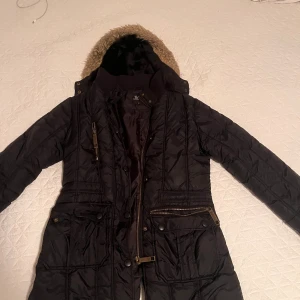 Svart quiltad parkajacka med fuskpäls - Svart lång parkajacka från Vero Moda med quiltad design, huva med fuskpäls och flera fickor med dragkedjor och knappar. Jackan har ribbad krage och är helfodrad för extra värme. Perfekt för kalla dagar och har en snygg urban vibe.