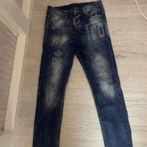 Dsquared2 mörkblå jeans med tryck - Snygga mörkblå jeans från Dsquared2 med coolt tryck på framsidan och slitna detaljer. Modellen har raka ben, klassisk femficksdesign och vita färgstänk för en edgy look. Jeansen har Dsquared2-logga på röd etikett vid fickan/ priset kan diskuteras 