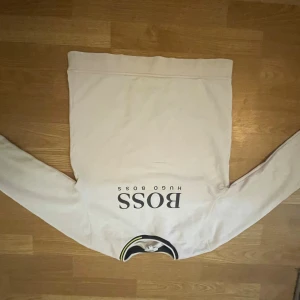Vit sweatshirt från Hugo Boss - Vit långärmad sweatshirt från Hugo Boss med svart och gul detalj vid kragen. Klassisk design med tryckt BOSS-logga på bröstet. Mjuk och skön, perfekt för dig som gillar stilrena plagg med snygga detaljer.