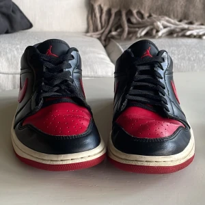 Nike Air Jordan 1 Low svart/röd EUR 39 - Nike Air Jordan 1 Low i svart och rött skinn med klassisk röd Swoosh och Air Jordan-logga broderad på hälen. Låg modell med snörning, rund tå och platt sula. Perfekt för dig som gillar ikoniska sneakers med streetkänsla.