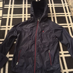 Mörkblå vindjacka från Polo Ralph Lauren - Snygg mörkblå vindjacka från Polo Ralph Lauren med huva och röda detaljer på dragkedjorna. Jackan har två fickor med dragkedja och klassisk Polo-logga broderad på bröstet. Perfekt för blåsiga dagar och enkel att matcha med streetwear.