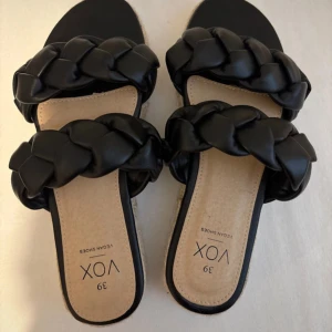 Svarta flätade sandaler från VOX - Säljer ett par svarta sandaler från VOX med två breda, flätade remmar i syntetmaterial. Sulan är platt med en snygg jutedetalj på kanten och ljus innersula. Perfekt för dig som gillar trendiga och bekväma skor med lite extra attityd.