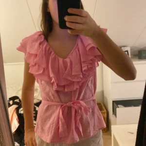 Rosa volangblus Holly & Whyte/ Lindex  - Supersöt rosa blus från Holly & Whyte med volanger runt v-ringningen och axlarna. Blusen är ärmlös och har ett knytband i midjan som kan tas av. Perfekt för dig som gillar romantiska detaljer och en luftig känsla. PASSAR MIG SOM HAR XS/S, använd 1 gång!!!!