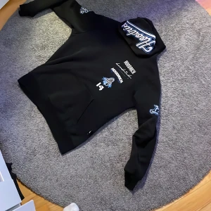 Svart hoodie från Hoodrich med tryck - Svart hoodie från Hoodrich med stora broderade och tryckta detaljer i vitt och blått på bröst, rygg, ärmar och huva. Hoodien har känguruficka, huva med tryck och coola street-inspirerade motiv. Perfekt för dig som gillar streetwear och vill sticka ut. Har små fickor på sidan som inte syns som gör det mycket lättare att hålla till exempel AirPods