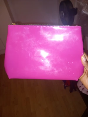 Rosa YSL kuvertväska i syntet - Snygg och stilren kuvertväska från YSL i blankt rosa syntetmaterial. Väskan har guldigt blixtlås och präglad YSL-logo framtill. Perfekt att bära i handen när du vill sticka ut med en färgklick.