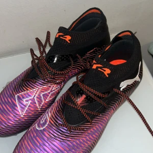 Puma Future 8 Ultimate FG fotbollsskor - Säljer ett par Puma Future 8 Ultimate FG fotbollsskor i svart, lila och orange med coola detaljer och snörning. Skorna har en syntetisk ovandel med mönstrad yta och Puma-logga på sidan. Perfekta för spel på fasta underlag.