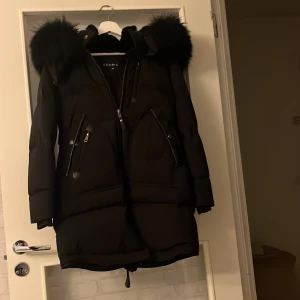 Svart pufferjacka med päls från Cedric - Svart pufferjacka från Cedric i storlek XS med fluffig päls på huvan, flera dragkedjor och stora fickor framtill. Jackan har lång ärm, snygg passform och är perfekt för kalla dagar. Snygg och stilren look med detaljer som dragsko nedtill och markerad huva. Pris kan diskuteras 