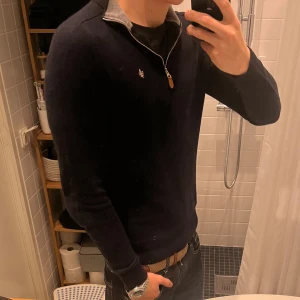 Marinblå merinoullströja Morris M - Snygg marinblå tröja från Morris i 100% merinoull. Tröjan har half zip med läderdetalj på dragkedjan och en broderad logga på bröstet. Långärmad och ribbad vid muddar och nederkant, perfekt för dig som gillar stilrena och klassiska plagg.