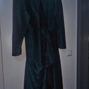 Glittrig långklänning från Shein Curve - Mörkgrön glittrig långklänning från Shein Curve i storlek 2XL. Klänningen har långa ärmar och är tillverkad i mjuk polyester. Snygg drapering framtill och skimrande material som fångar ljuset. Perfekt för dig som vill sticka ut med stil.