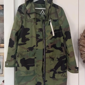 Camouflage regnjacka WearColour XS - Snygg regnjacka från WearColour i storlek XS med klassiskt camouflage-mönster i gröna och svarta toner. Jackan har huva, stora fickor framtill och dragkedja med tryckknappar. Perfekt för blöta dagar och outdoor-häng.