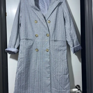 Rutig kappa från NLY Trend, strl 36 - Snygg rutig kappa från NLY Trend i ljusgrå toner, dubbelknäppt med stora beige knappar och klassisk krage. Jackan har långa ärmar och två fickor framtill. Perfekt för dig som gillar stilrena och trendiga ytterplagg.