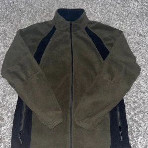 Mörkgrön fleecejacka med svarta detaljer - Snygg mörkgrön fleecejacka med svarta paneler längs sidorna och ärmarna. Jackan har hel dragkedja framtill, högt krage och två sidofickor med dragkedja. Perfekt för kyliga dagar och enkel att matcha med jeans eller mjukis.