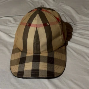 Burberry rutig keps i beige och svart - Snygg Burberry keps med klassiskt rutigt mönster i beige, svart och röda detaljer. Kepsen har böjd skärm och justerbar passform. Tillverkad i bomull, perfekt för dig som vill ha en ikonisk streetstil med lyxig touch.