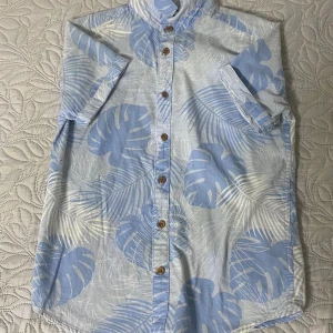 Blå kortärmad skjorta från Kappahl - Fräsch kortärmad skjorta från Kappahl med ljusblå och vita tropiska bladmönster. Skjortan har träfärgade knappar framtill och klassisk krage. Tillverkad i mjuk bomull, perfekt för varma dagar och chill stil.