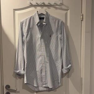 Blå och vit Randig skjorta från Ralph Lauren XS - Snygg skjorta från Ralph Lauren i storlek XS med k blåvitt randigt mönster. Skjortan har knappar framtill, button-down krage och  broderade loggan på bröstet. Har långa ärmar med manschettknappar.  Passar även S/M. Jag är 184