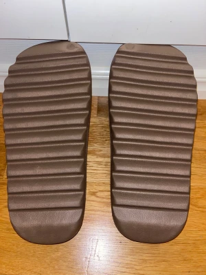 Adidas bruna slides med räfflad sula - Säljer ett par bruna Adidas slides med chunky, räfflad sula och stilren design. De har en bred rem över foten och är tillverkade i syntetmaterial för maximal komfort. Perfekta för chill dagar eller att glida runt hemma. Adidas-loggan är diskret präglad på fotbädden.