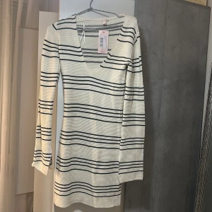 Randig stickad klänning från Nelly - Superfin stickad klänning från Nelly i vitt med svarta ränder. Klänningen har lång ärm, v-ringning och en kort passform som sitter snyggt på kroppen. Perfekt för dig som gillar en enkel men stilren look.