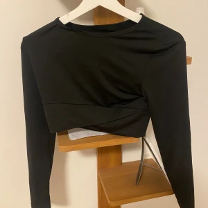 Svart croppad topp från SHEIN - Svart långärmad croppad topp från SHEIN med rund halsringning och stilren twistad detalj framtill. Tillverkad i stretchigt material som sitter snyggt på kroppen. Perfekt för dig som gillar en clean och modern look. Funkar perfekt till gymmet också.