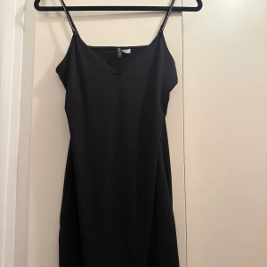 Svart klänning med glittriga axelband H&M - En svart kortklänning från H&M med smala axelband dekorerade med glittriga stenar. Klänningen har v-ringning och en enkel, stilren design som ger en snygg och trendig look. Perfekt för dig som gillar minimalistiskt men ändå vill ha en touch av glitter.