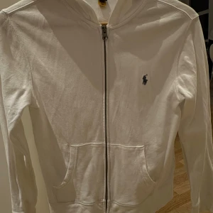 Vit hoodie från Polo Ralph Lauren - Vit hoodie från Polo Ralph Lauren med dragkedja framtill och klassisk blå logga på bröstet. Tröjan har huva, två fickor och är tillverkad i mjuk bomull. Perfekt för chill dagar och enkel att matcha med jeans eller joggers.