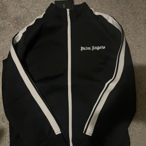 Palm angels kofta  - Palm Angels svart trackjacka med vita ränder längs ärmarna och vit dragkedja framtill. Jackan har hög krage och logotypen broderad på bröstet. Tillverkad i mjuk polyester som ger en sportig och clean vibe. Perfekt för streetwear-stilen.