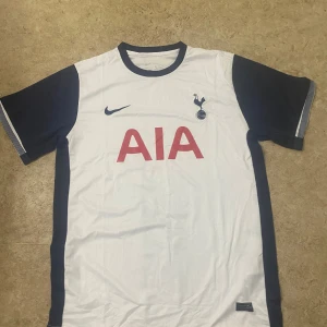 Tottenham Hotspur vit/blå matchtröja - Säljer en Tottenham Hotspur fotbollströja från Nike med AIA-tryck på bröstet. Tröjan är vit med mörkblå ärmar och detaljer, har klubbens emblem på vänster sida och Nike-logga på höger. Tillverkad i lätt och ventilerande material, perfekt för träning eller match.