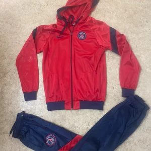 PSG träningsset röd och blå Nike - Snyggt PSG träningsset från Nike med röd huvtröja och blå byxor. Jackan har dragkedja, huva med snörning och PSG-logga på bröstet. Byxorna är mörkblå med PSG-emblem och har resår i midjan. Perfekt för dig som älskar fotboll och vill ha en sportig look.