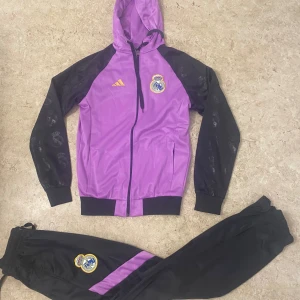 Real Madrid lila träningsset Adidas - Snyggt träningsset från Adidas x Real Madrid med lila huvtröja och svarta ärmar med Real Madrid-loggor. Matchande svarta byxor med lila detalj och broderad klubbmärke. Jackan har dragkedja och huva, byxorna har resår och snörning i midjan.